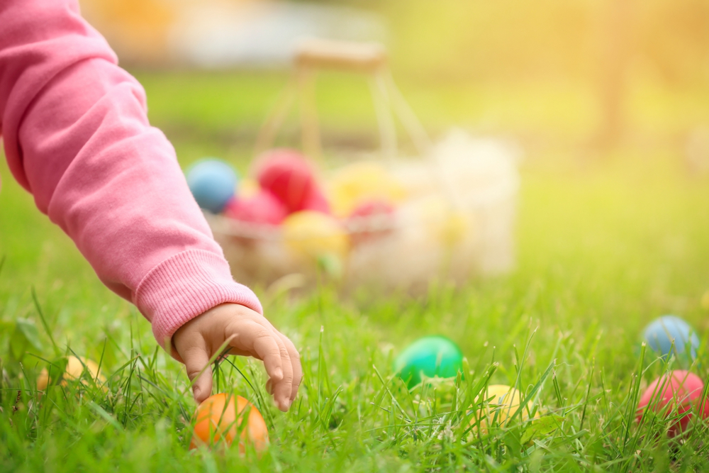 Featured image showing a child's hand picking up an easter egg in tall green grass during an Easter Egg Hunt.